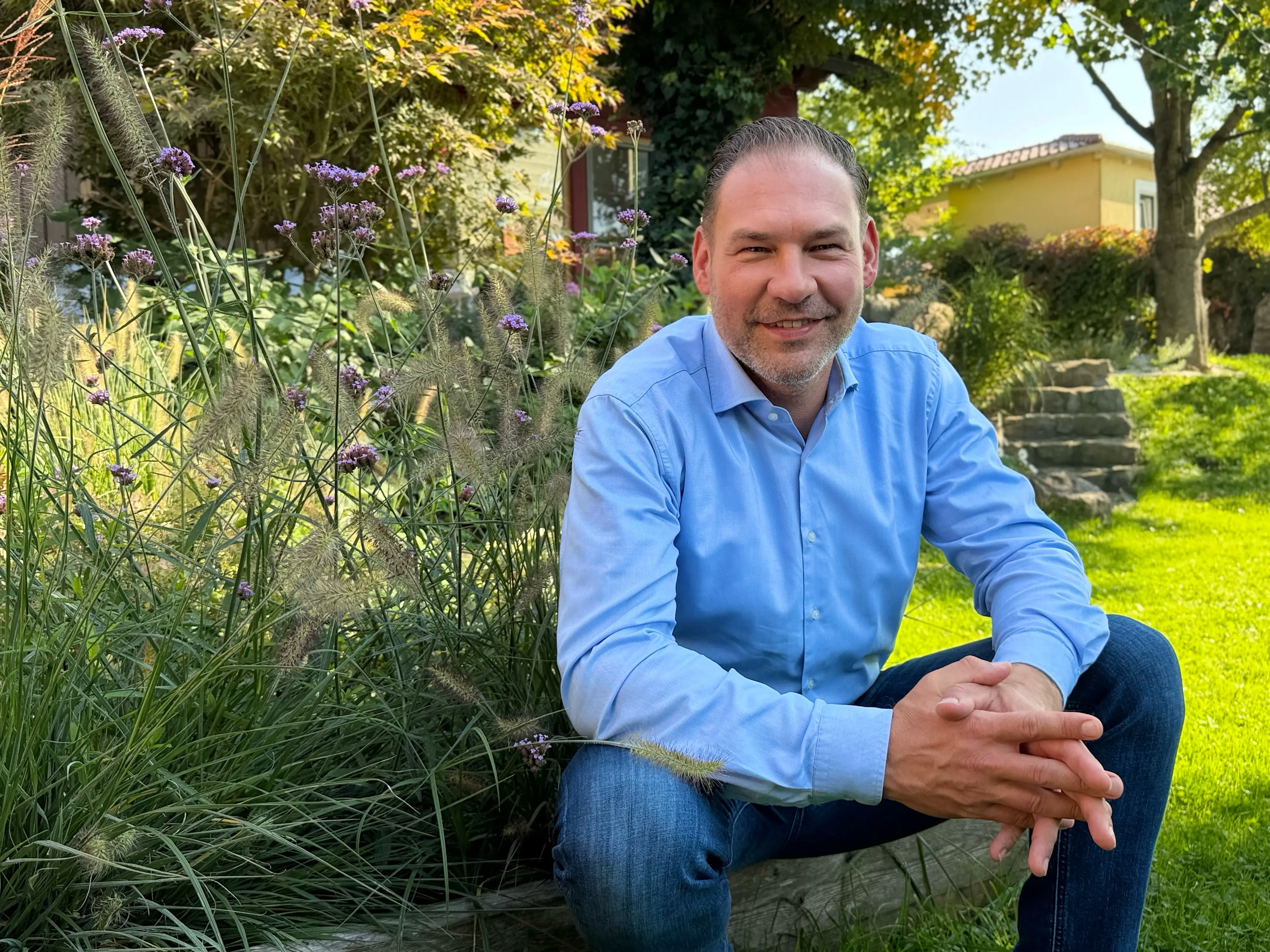 Christian Görisch sitzt im Hemd in einem Garten zwischen hohen Gräsern